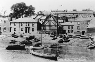There are some two dozen rowboats moored just off the beach or drawn up on land in the foreground. Behind them, what appears to be a terrace of two-storey houses is to the left of the scene, the gable end of the old half-timbered Mission Hall with its steeply-pitched roof is next, and the Union Inn can be seen to the right. Just visible from this low angle, behind and in the gaps between these buildings, are some of the houses in Tamar Street and on the hill behind.