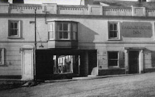 This close-up photo shows most of the front of the old Passage House Inn, with its second storey passing over the end of Tamar Street. Part of Tamar Street can be seen through the opening, which has a bay window above it. A doorway opens into the street to the right, below the arch, and there is another door in the right frontage, flanked on each side by a window. Above this door, between another pair of windows, the name of the Inn is painted. The whole front wall of the building is surmounted by a fancy balustrade which conceals the roof.