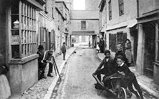 Looking along Tamar Street from the southern end towards the Passage House Inn, there is a group of three boys with a wooden wheelbarrow in the foreground. On the narrow cobbled pavement to the left, behind the bay window of a shop, an artist sits on a low stool with his easel in front of him. The buildings fronting on the narrow street are varied in appearance, with some being whitewashed, others bare brick, and at least one slate-fronted. At the far end, the upper storey of the Inn passes over the street, with a solitary window in the centre.